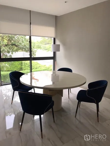 独立式住宅 出租在Watthana, Bangkok - 采光良好的餐区，白色圆桌与四把海军蓝椅，临大窗与百叶，地面为大理石。