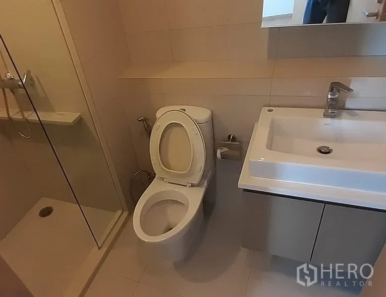 Condominium for rent in Khlong Toei, Bangkok - Contemporary bathroom with vanity sink, toilet, and a walk-in shower in neutral tiles.