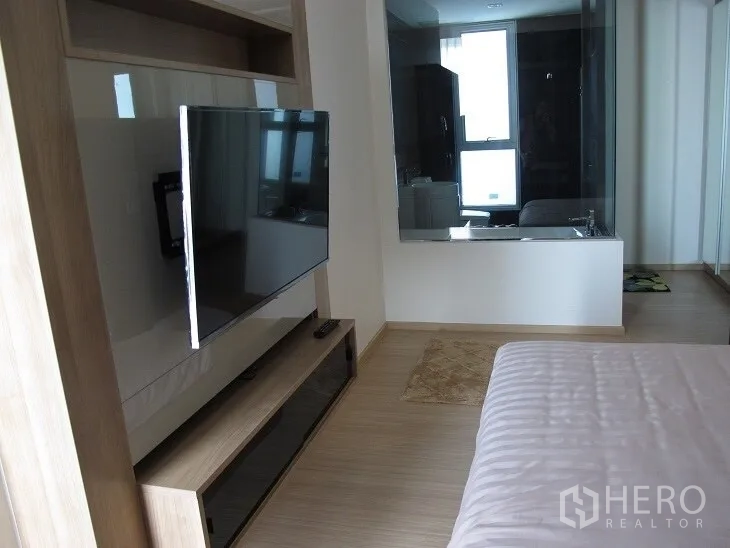 Condominium for rent or sale in Khlong Toei, Bangkok - Primary bedroom with wall-mounted TV and glass window to the bathroom over wood flooring.