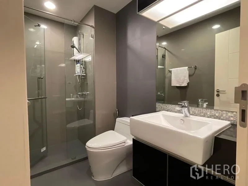 Condominium for sale in Khlong Toei, Bangkok - Modern bathroom with glass-enclosed shower, vanity sink and toilet.