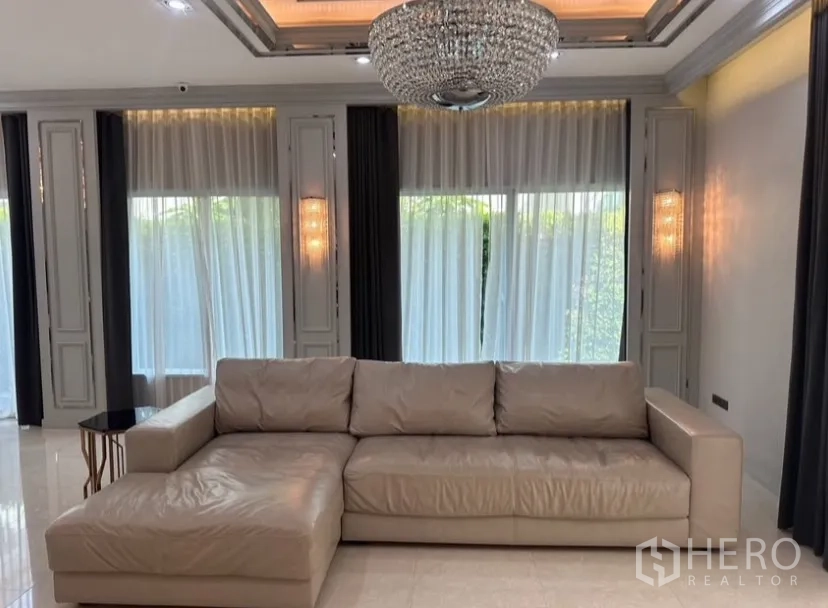 Detached House for rent or sale in Prawet, Bangkok - Ground-floor living area featuring an L-shaped leather sofa, crystal chandelier, and floor-to-ceiling windows.