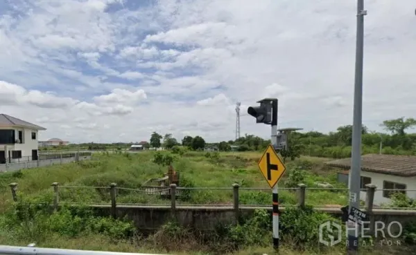 Land for sale in Bang Sao Thong, Samut Prakan - Roadside view of the plot with traffic sign, low fence and nearby houses across open fields.