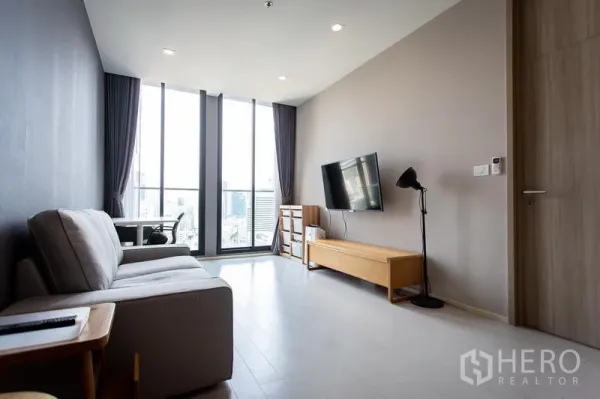 คอนโด ให้เช่าในPathum Wan, Bangkok - ห้องนั่งเล่นสว่างพร้อมหน้าต่างสูงจรดเพดาน วิวเมือง โซฟา และทีวีติดผนัง ในคอนโดปทุมวัน