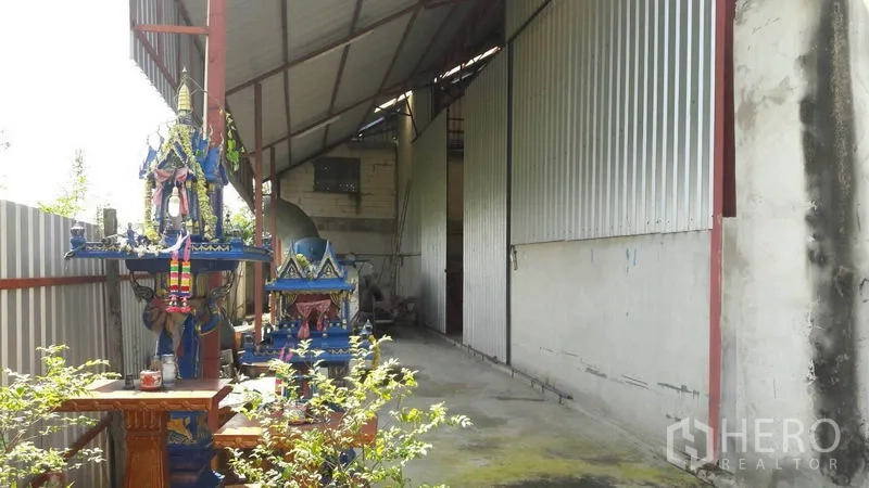 Warehouse for sale in Pathum Thani - Exterior corridor along corrugated metal walls with covered roof and outdoor area near the entrance.