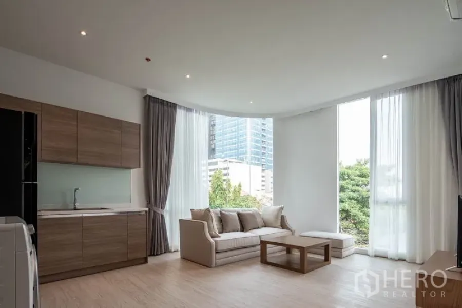 Condominium for rent in Khlong Toei, Bangkok - Bright open‑plan living room with kitchenette, sofa and floor‑to‑ceiling windows overlooking city greenery.