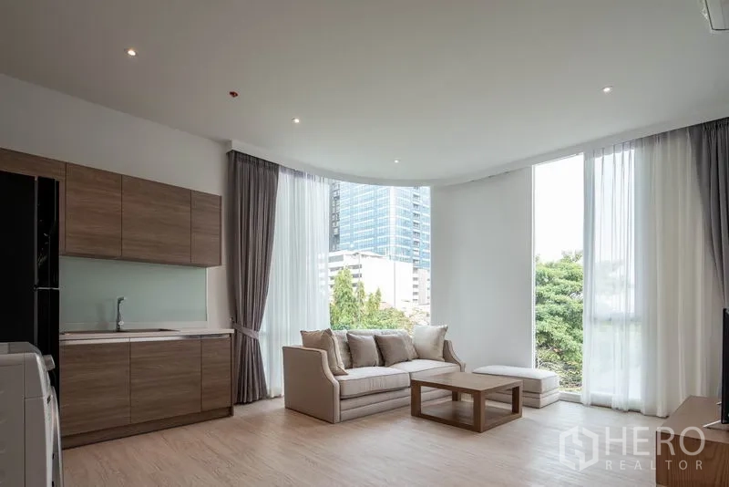 Condominium for rent in Khlong Toei, Bangkok - Bright open‑plan living room with kitchenette, sofa and floor‑to‑ceiling windows overlooking city greenery.