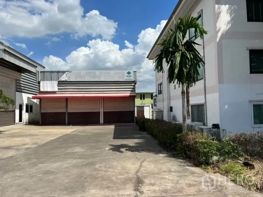 Warehouse for rent in Prawet, Bangkok - Front view of a warehouse with wide concrete yard, red canopy and multiple roller shutters beside an office building and palm tree under a blue sky.