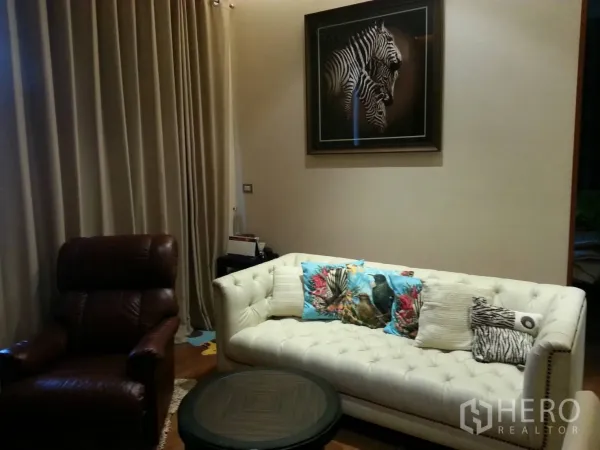 Condominium for rent or sale in Khlong Toei, Bangkok - Cozy living room with white tufted sofa, brown recliner, round coffee table, zebra artwork and full-length curtains.