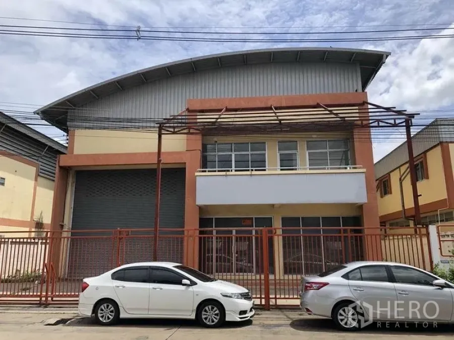 โรงงาน ให้เช่าในPathum Thani - อาคารโรงงานสองชั้น หลังคาเมทัลชีททรงโค้ง ประตูม้วนขนาดใหญ่ ด้านหน้าเป็นออฟฟิศกระจก มีรั้วรอบและที่จอดรถริมถนน