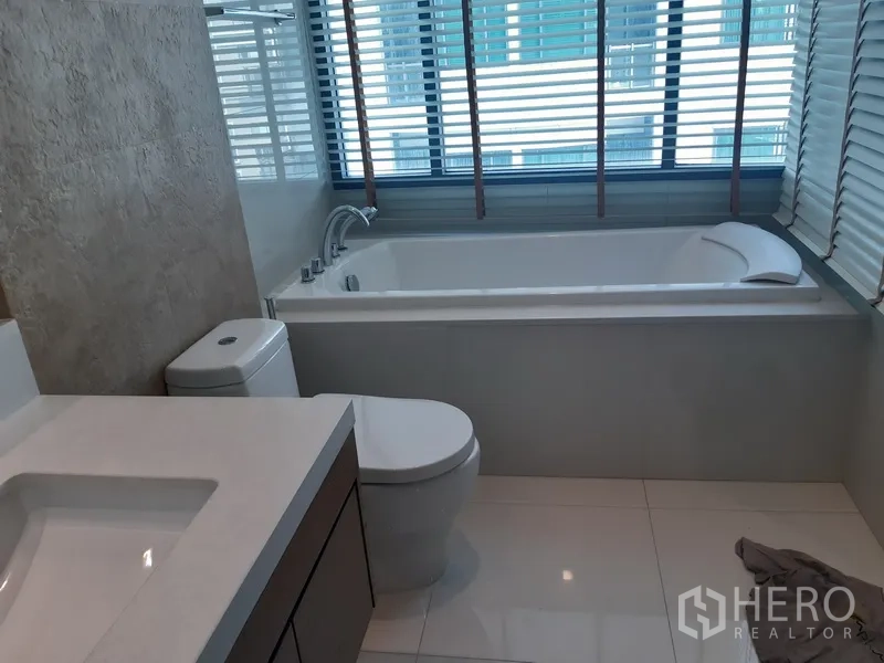 Condominium for rent or sale in Khlong Toei, Bangkok - Contemporary bathroom featuring a large soaking bathtub by the window, toilet and modern vanity.