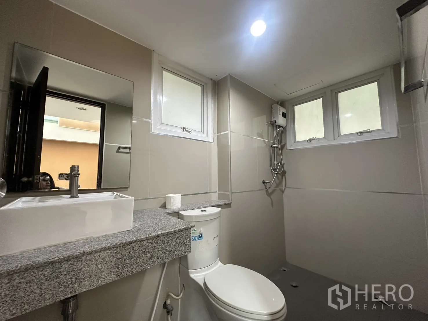 โฮมออฟฟิศ for rent in Watthana, Bangkok - ห้องน้ำปูกระเบื้องโทนเทา มีอ่างล้างหน้า กระจก สุขภัณฑ์ และฝักบัวพร้อมเครื่องทำน้ำอุ่นใต้หน้าต่างบานเล็ก