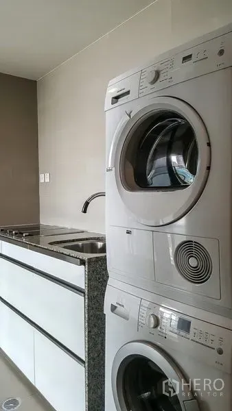 Condominium for rent or sale in Sathon, Bangkok - Laundry area with stacked washer and dryer beside a double sink and storage cabinets.