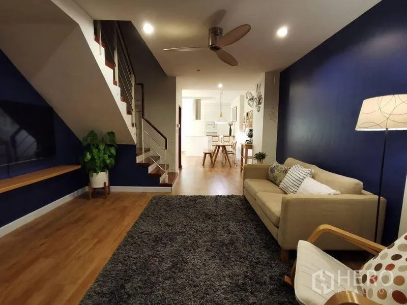 Townhouse for rent or sale in Yan Nawa, Bangkok - Open-plan living room with staircase, navy accent wall, sofa, ceiling fan, rug and wall-mounted TV.