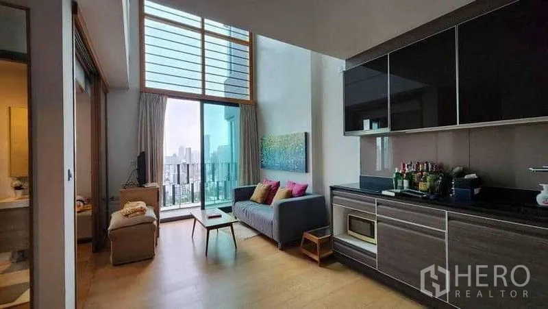 Condominium for rent in Khlong Toei, Bangkok - Double-height living room in Khlong Toei condo with balcony and skyline view, open kitchenette.