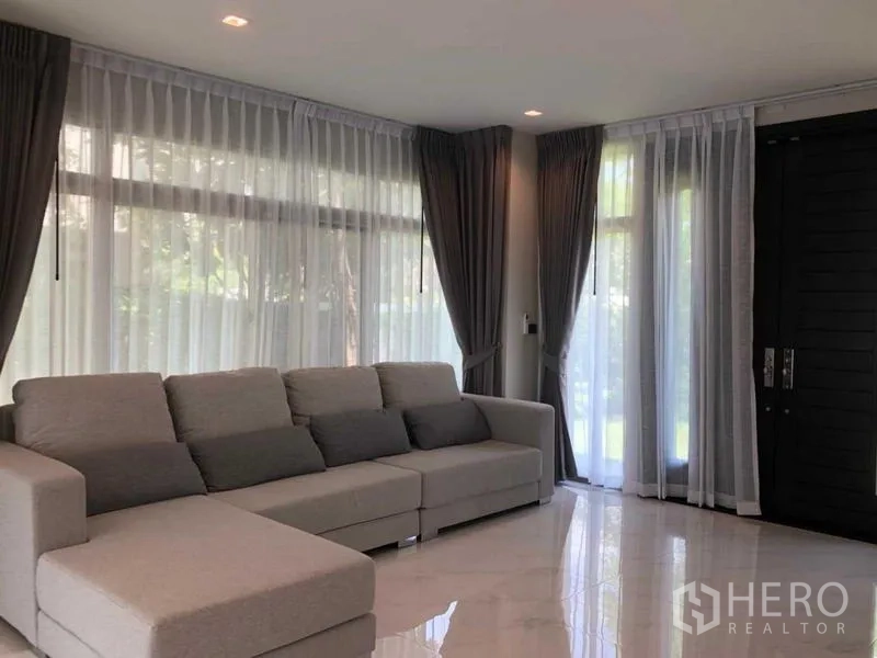 Detached House for rent in Lat Krabang, Bangkok - Corner living room featuring an L-shaped sofa and floor-to-ceiling windows with sheer drapes.