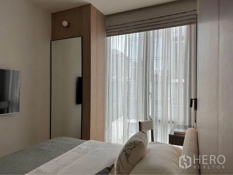Condominium for rent in Pathum Wan, Bangkok - Bedroom looking to a tall window with sheer curtains, slim mirror and wall-mounted TV.