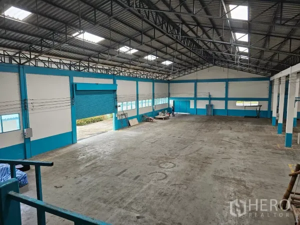 Factory for rent or sale in Mueang Rayong, Rayong - Spacious factory interior with high steel truss roof, skylights, heavy-duty concrete floor and a large blue roller shutter door.