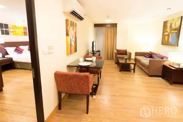 Condominium for rent in Khlong Toei, Bangkok - Open living–dining area with two-seat table, wall art and a doorway to the bedroom with warm lighting.