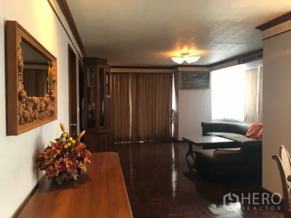 Condominium for rent in Khlong Toei, Bangkok - Warm living room with parquet flooring, L-shaped sofa, wooden cabinetry and curtained windows.
