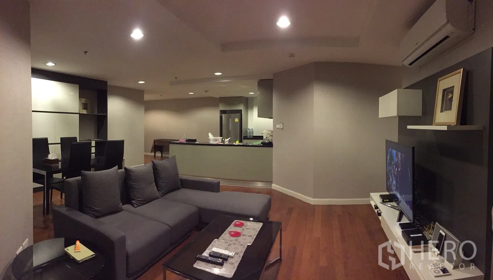 คอนโด ให้เช่าในHuai Khwang, Bangkok - ห้องนั่งเล่นแบบโอเพ่นแพลน โซฟาสีเทา ผนังทีวี และพื้นไม้โทนอุ่น