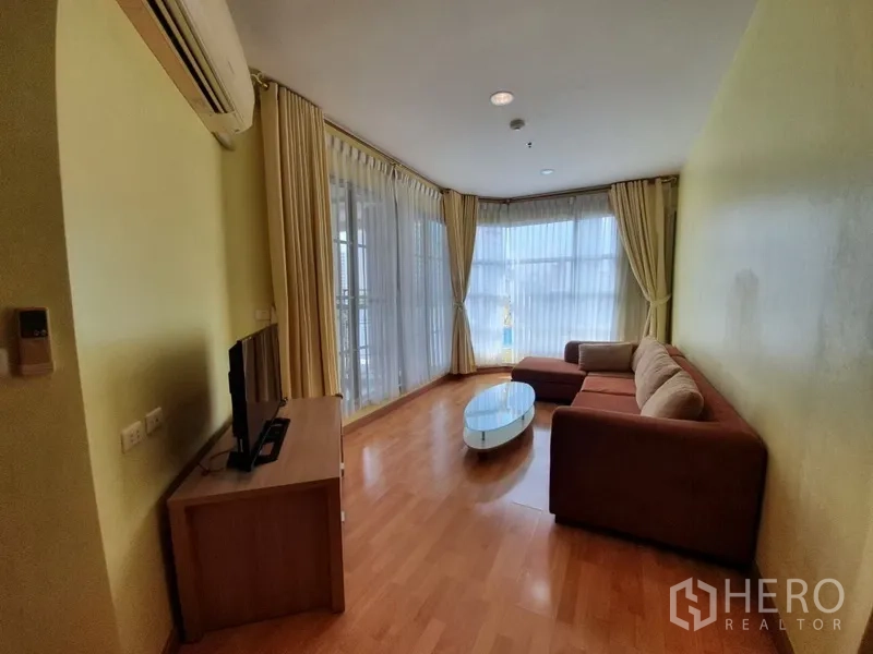 Condominium for rent in Khlong Toei, Bangkok - Bright corner living room with floor-to-ceiling windows, L-shaped sofa, TV console and wood flooring.