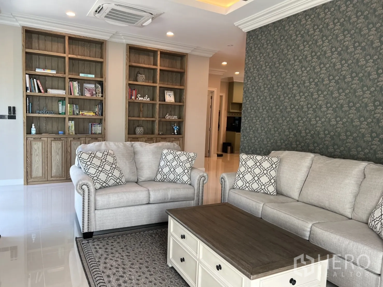 Detached House for rent or sale in Bang Kapi, Bangkok - Bright living room featuring built-in wooden bookshelves, patterned wallpaper, and a set of light-gray sofas around a coffee table.