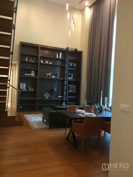 Condominium for rent in Khlong Toei, Bangkok - Loft-style dining area with high ceiling, staircase, pendant lighting, and full-height shelving.