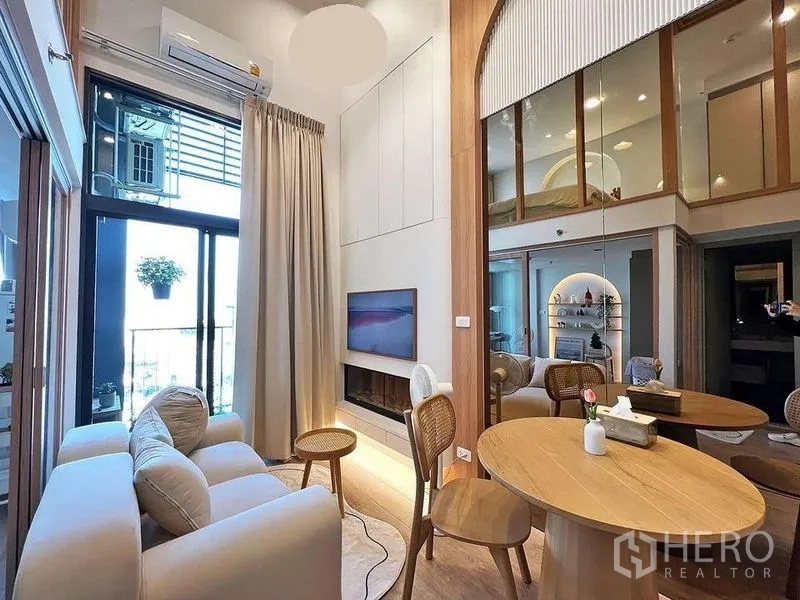 Condominium for rent in Huai Khwang, Bangkok - Double-height living room with balcony doors, mounted TV, neutral sofa and round dining table.