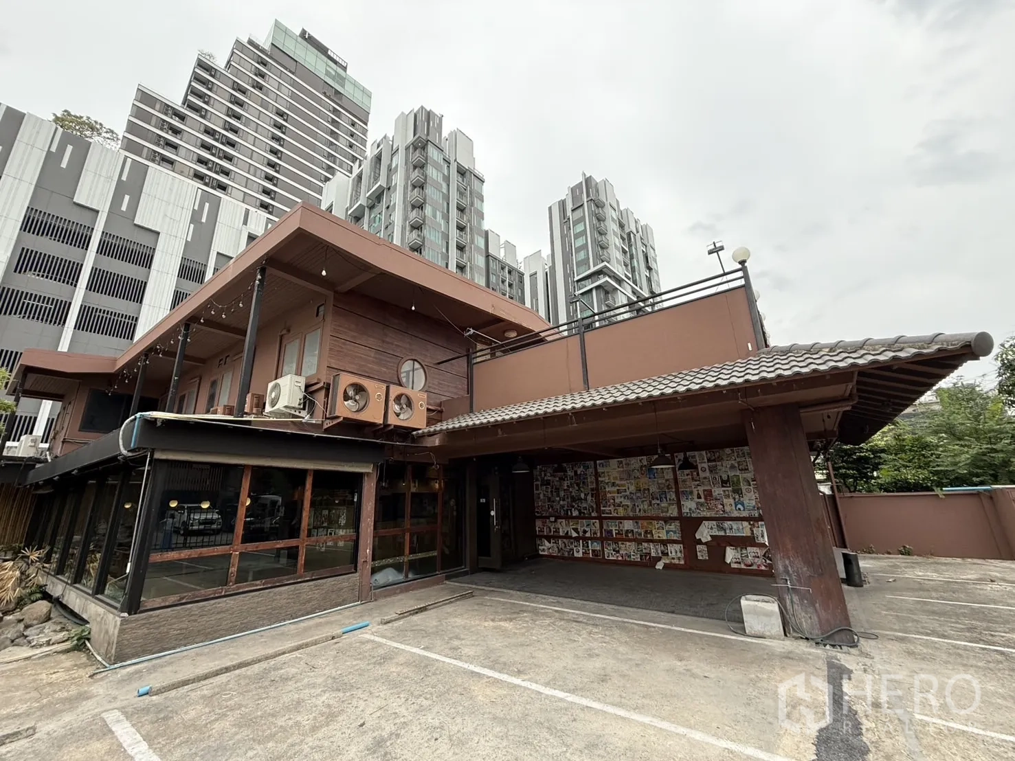 Detached House for rent in Watthana, Bangkok - Two-storey detached house with wood accents and covered parking, set against nearby high-rise buildings near Ekkamai.