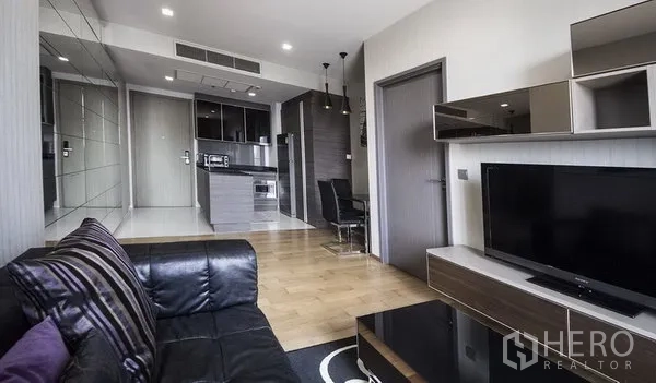 Condominium for rent in Khlong Toei, Bangkok - Open-plan living room with leather sofa, TV console, and compact kitchen with dining in a Sukhumvit condo.