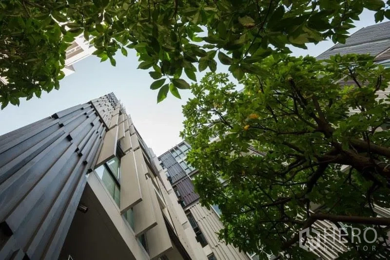 公寓 出租在Watthana, Bangkok - 现代公寓楼外观仰视图，绿树环绕。