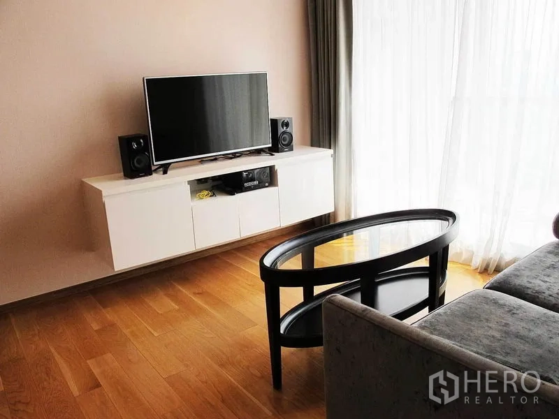 Condominium for rent in Watthana, Bangkok - Bright living room with floating TV cabinet, flat-screen TV, speakers, oval glass coffee table and full-height window with sheer curtains.