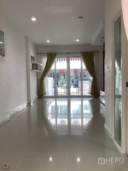 Townhouse for rent or sale in Wang Thonglang, Bangkok - Bright living room with glossy tile floors, recessed lights, and sliding glass doors with green curtains opening to a small terrace.