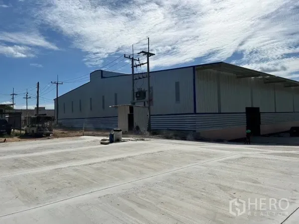 Warehouse for rent in Nikhom Phatthana, Rayong - Metal-clad warehouse exterior with large concrete yard and overhead power lines under a blue sky in Nikhom Phatthana.