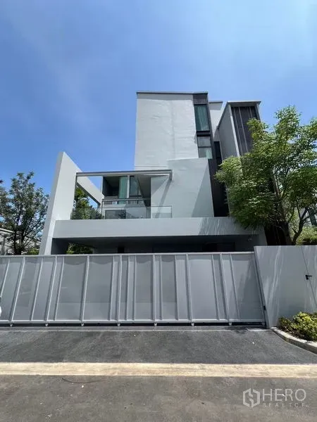 Detached House for rent in Saphan Sung, Bangkok - Modern three-storey detached house exterior with tall privacy fence and trees under a blue sky.