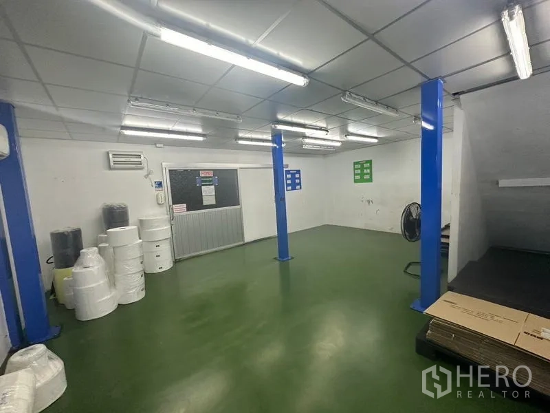 Warehouse for rent or sale in Lam Luk Ka, Pathum Thani - Open work area with blue steel columns, rolls of packing material and epoxy floor.