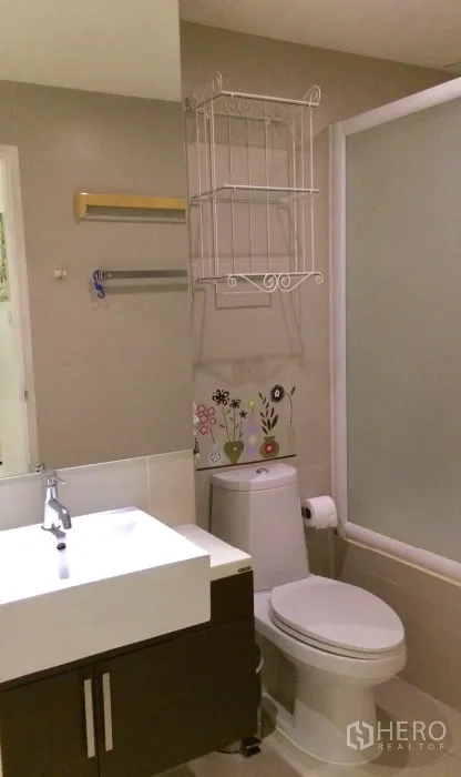 Condominium for rent in Khlong Toei, Bangkok - Modern bathroom with vanity sink, toilet and bathtub with frosted glass screen and decorative metal shelving.