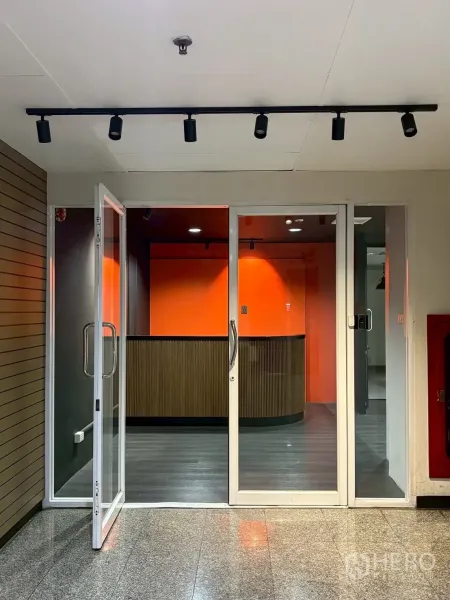 ออฟฟิศ for rent in Khlong Toei, Bangkok - ประตูกระจกหน้าออฟฟิศพร้อมไฟรางและเคาน์เตอร์ต้อนรับผนังสีส้ม