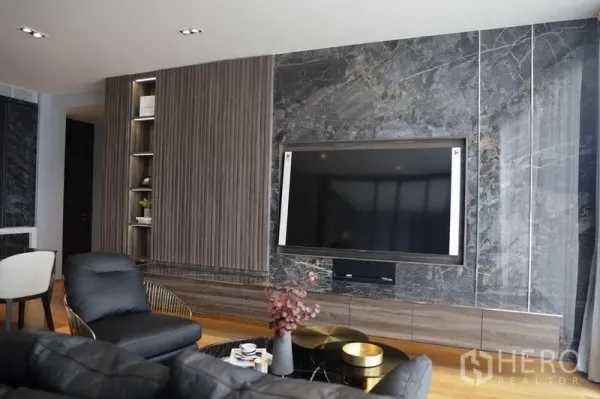 Condominium for rent in Khlong Toei, Bangkok - Contemporary living room with a large TV mounted on a marble wall, built-in shelves, leather sofa and wood floors.