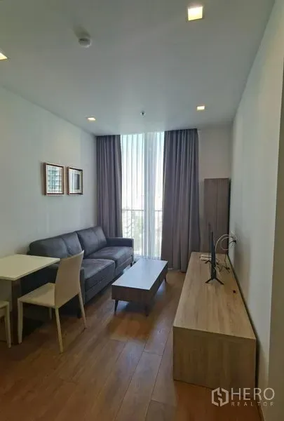 公寓 出租在Watthana, Bangkok - 明亮客厅，灰色沙发、电视柜与小餐桌，木地板，大落地窗配窗帘。