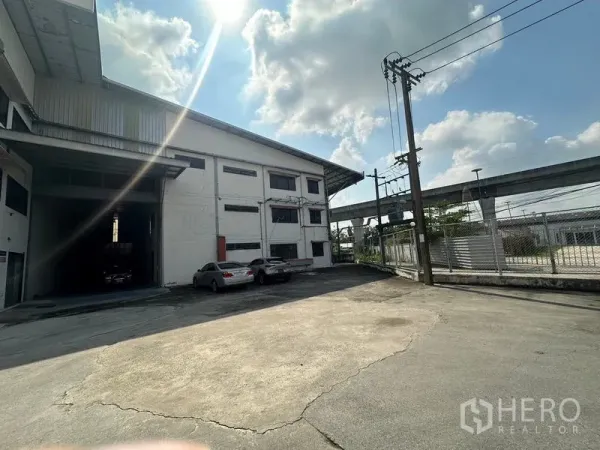 Warehouse for rent in Suan Luang, Bangkok - Front yard and gated entrance of the warehouse with power poles and parking area.