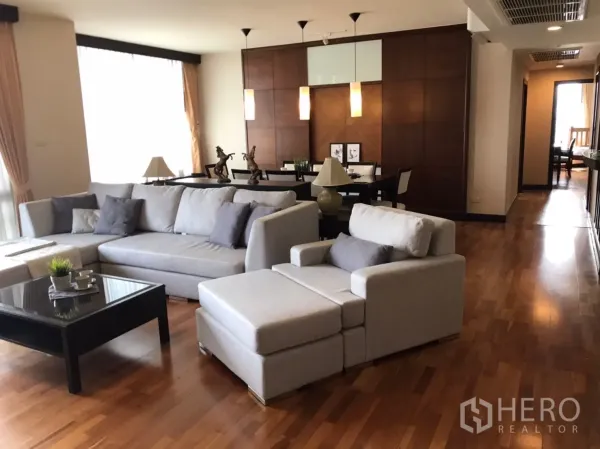 公寓 出租或出售在Pathum Wan, Bangkok - 采光良好的客厅，灰色转角沙发、黑色茶几、木地板，后方为吊灯餐区。