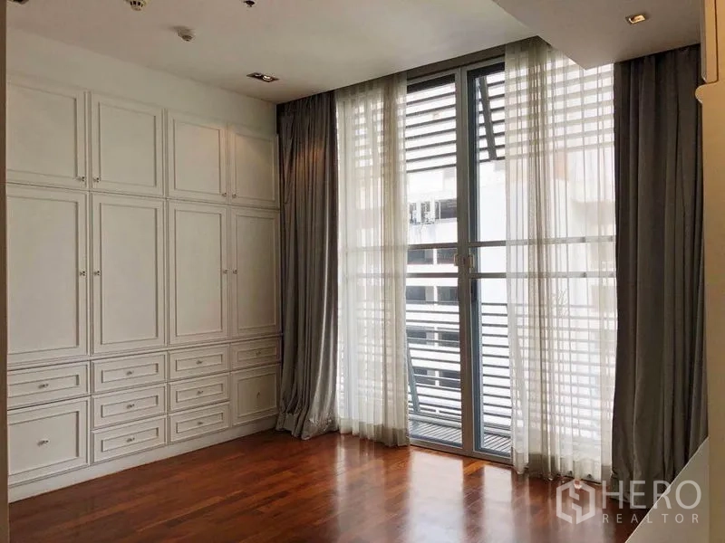 Condominium for rent in Khlong Toei, Bangkok - Room with floor-to-ceiling windows, sheer curtains and a full wall of white built-in cabinets.