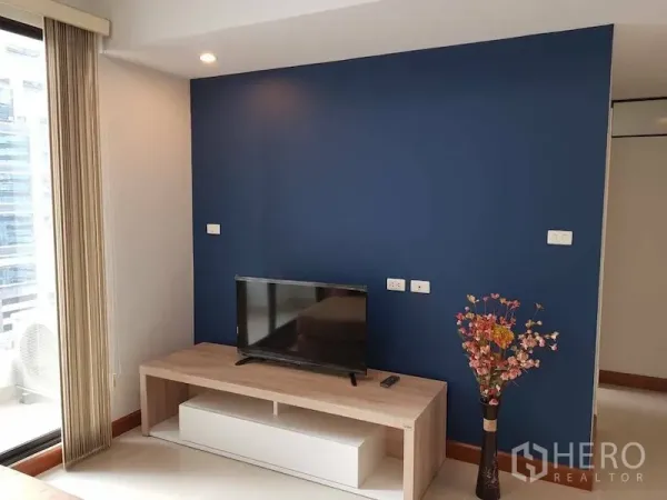 公寓 出租在Watthana, Bangkok - 海军蓝特色墙与木质电视柜和花瓶，旁有落地门通往阳台。