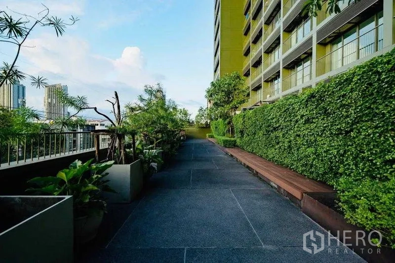 Condominium for rent in Watthana, Bangkok - Landscaped outdoor walkway with hedges, benches, and views toward the city skyline.
