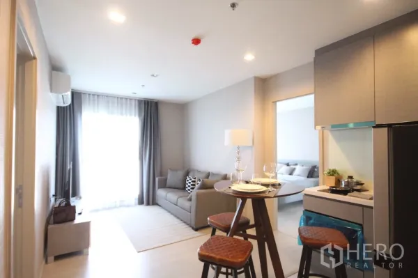 Condominium for rent in Khlong Toei, Bangkok - Bright open-plan living room with grey sofa, round dining table and compact kitchenette, facing a large curtained window.