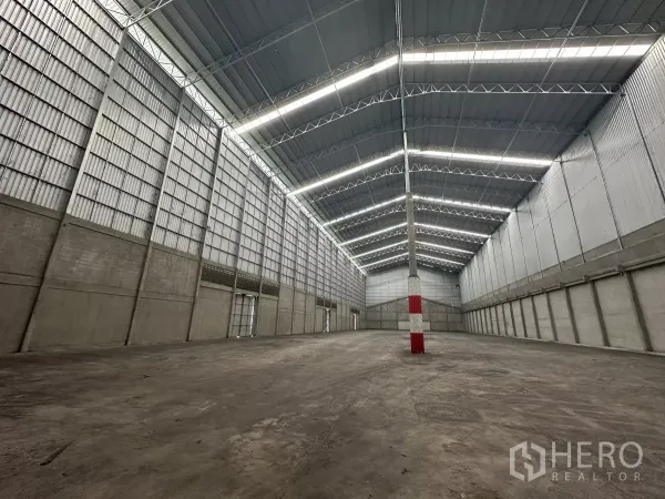 Warehouse for rent in Mueang Pathum Thani, Pathum Thani - Open storage hall with central red‑and‑white safety column, metal cladding walls and bright roof lighting.