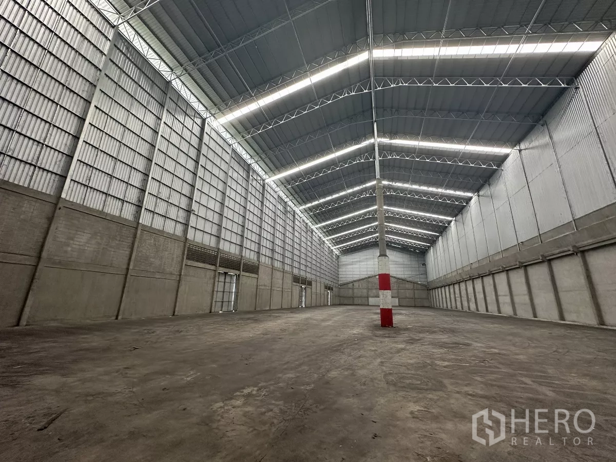 Warehouse for rent in Mueang Pathum Thani, Pathum Thani - Open storage hall with central red‑and‑white safety column, metal cladding walls and bright roof lighting.