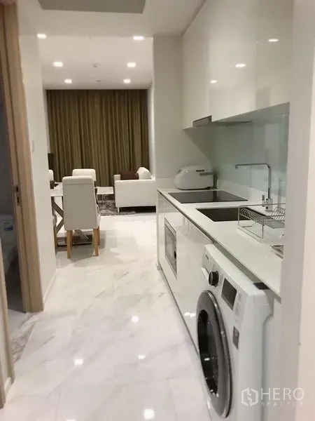 Condominium for rent in Watthana, Bangkok - Bright galley kitchen with white cabinetry and built‑in washer leading to the living–dining area.