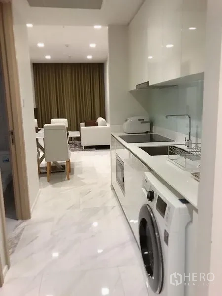 Condominium for rent in Watthana, Bangkok - Bright galley kitchen with white cabinetry and built‑in washer leading to the living–dining area.